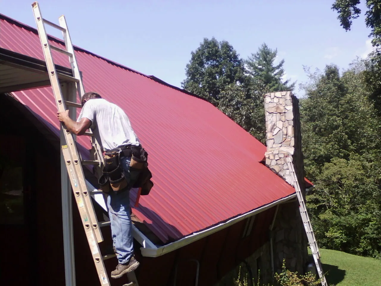 Roofer on a ladder beside a red metal roof and stone chimney — Soffit & Fascia Repair by Roofing Brothers in Lakehurst